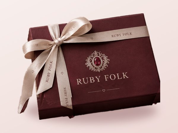 Ruby Folk gift box: burgundy lid with crest and wordmark, tied with a champagne ribbon